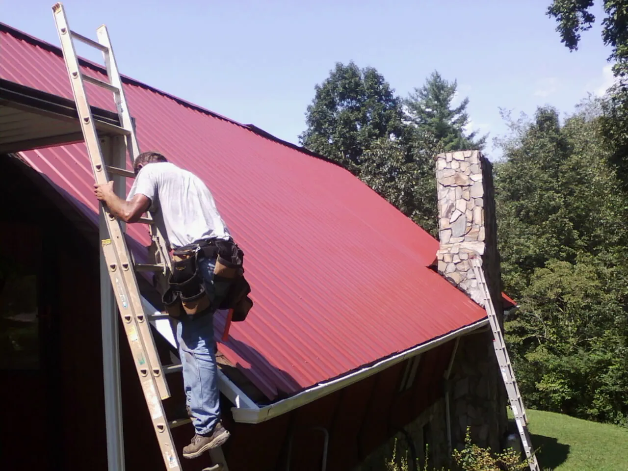 Roofer on a ladder beside a red metal roof and stone chimney — Soffit & Fascia Repair by Roofing Brothers in Cantrall
